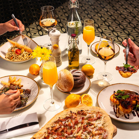 Mesa servida con platos de pasta, carne, focaccia y copas de vino blanco y jugo de naranja.