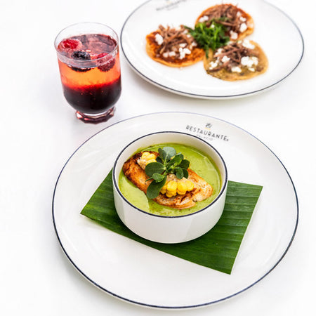 Tazón de crema verde con pollo y elote sobre hoja de plátano junto a sopes y bebida roja.