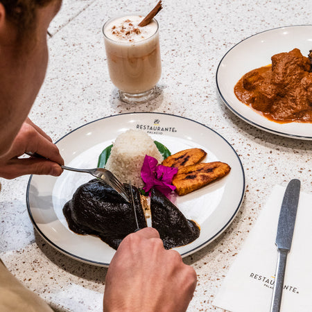 Pechuga de pollo bañada en mole oscuro con arroz blanco, plátanos fritos y pétalos morados sobre plato blanco.